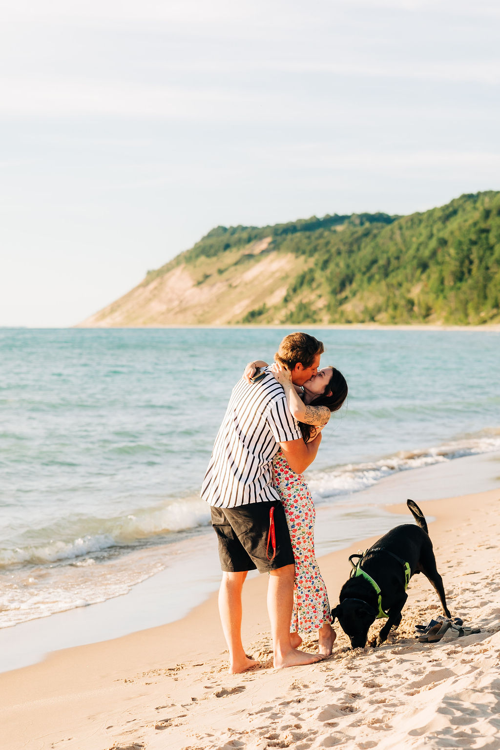 newly engaged couple kissing on beach with dunes in background as dog digs in the sand