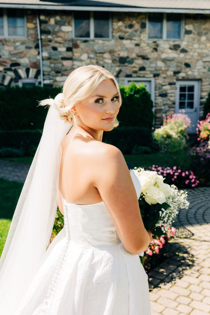 blonde woman wearing wedding dress and holding bouquet of flowers in front of brick cobblestone building